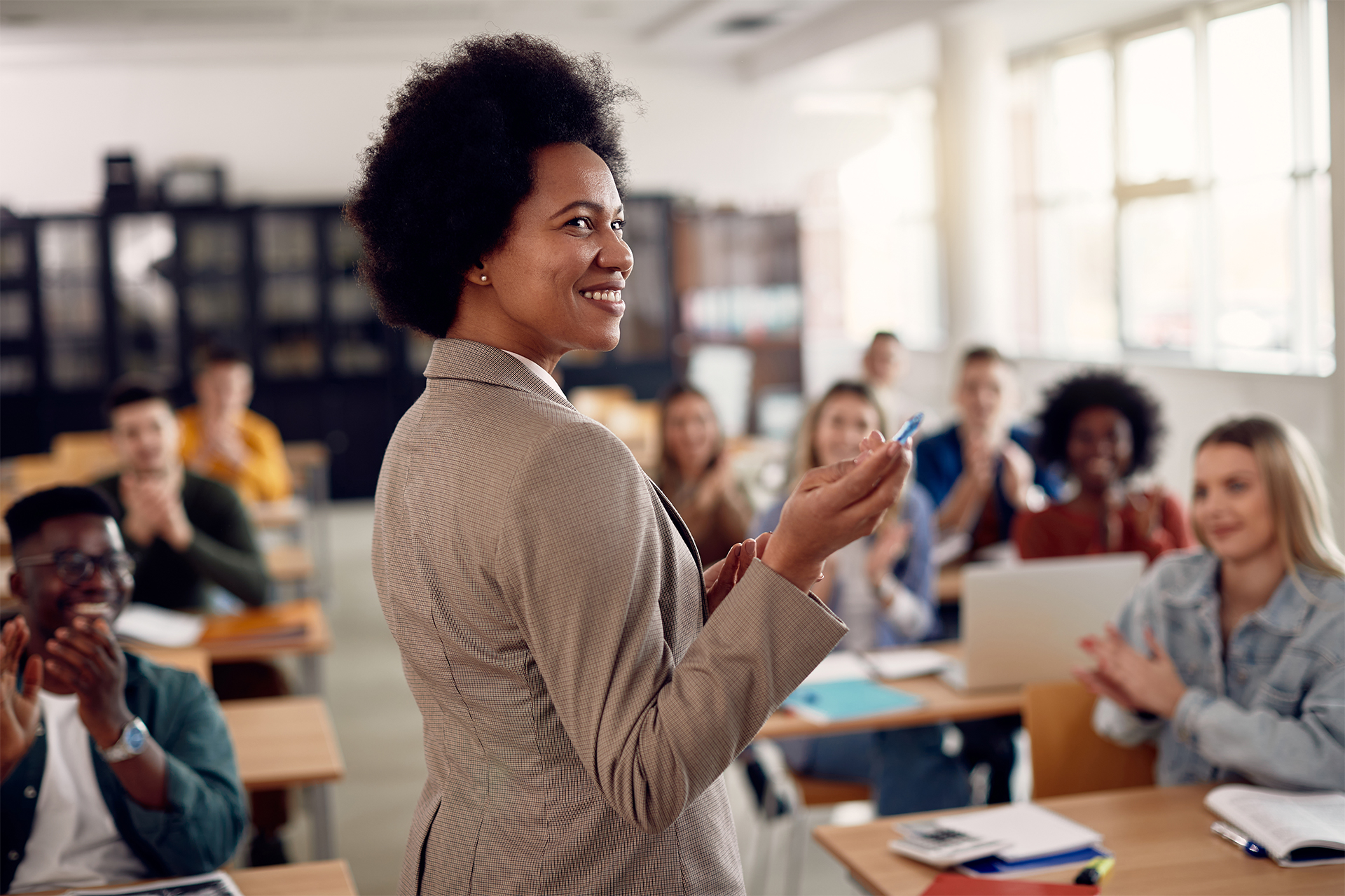 Satisfied professor stands at the front of her classroom as students smile and applaud her lecture.